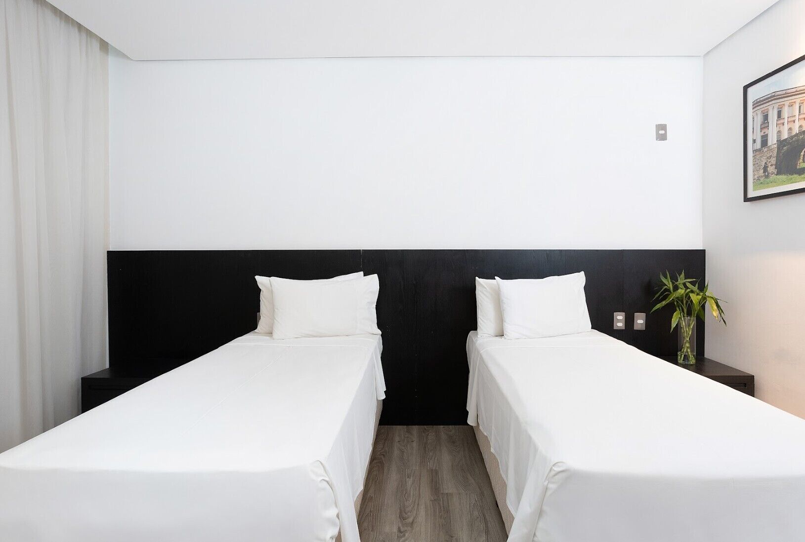 Quarto de hotel com duas camas de solteiro, cabeceira preta, mesa de cabeceira pequena com um vaso de plantas e um quadro na parede.
