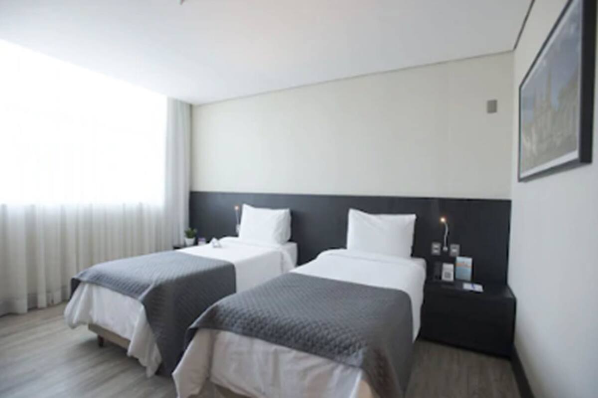 Habitación de hotel con dos camas, cabecera oscura, mesita de noche con lámpara y ventana con cortinas.