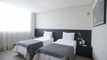 Habitación de hotel con dos camas, cabecera oscura, mesita de noche con lámpara y ventana con cortinas.