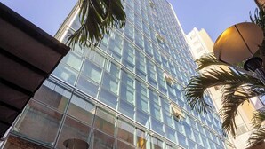 A modern glass building with a reflective facade, surrounded by palm trees and outdoor seating areas. - Hotel Park Tower Savassi (Belo Horizonte)
