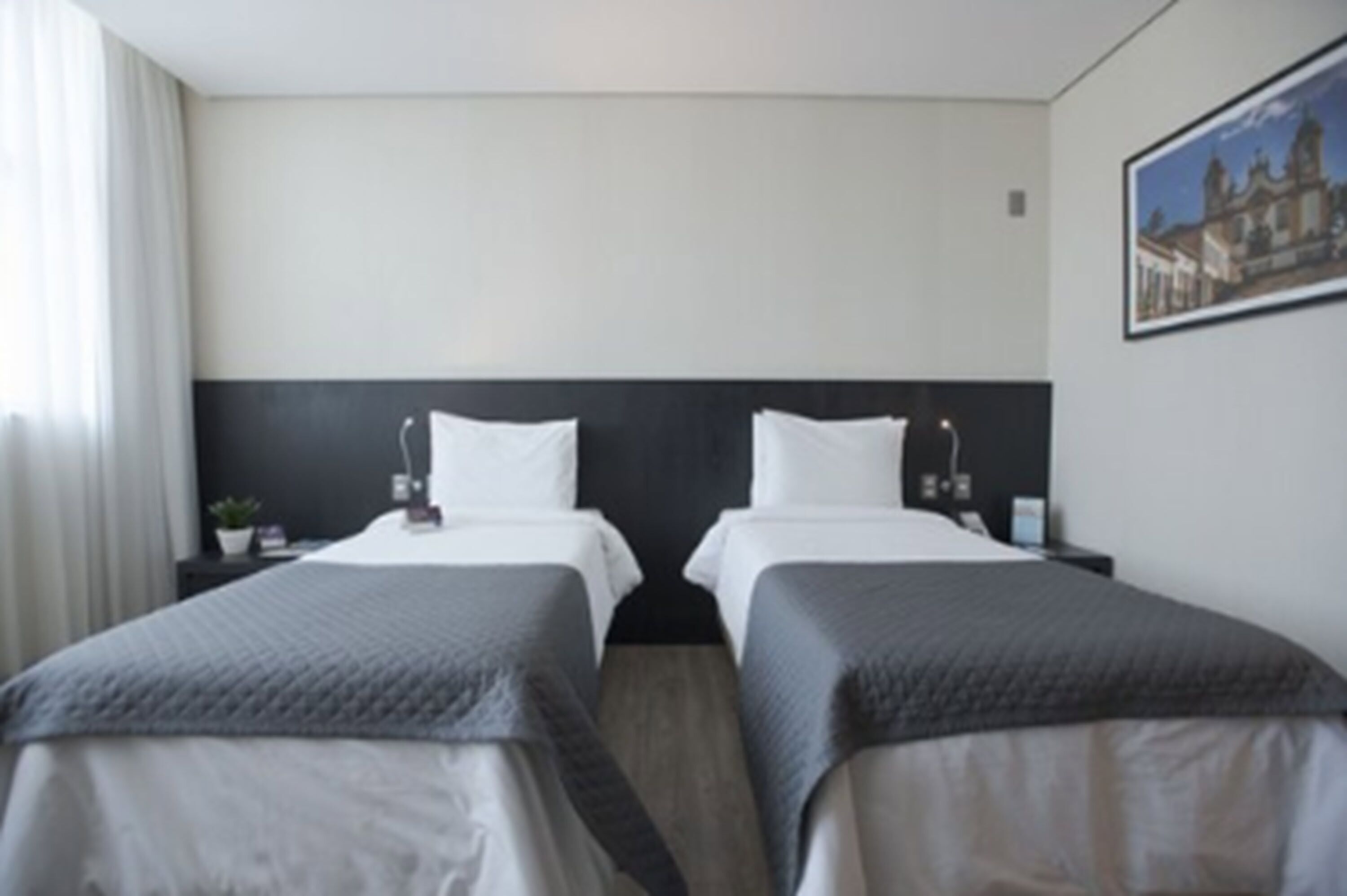 A hotel room with two beds, a nightstand with a lamp, a small table with a plant, and a framed picture on the wall.
