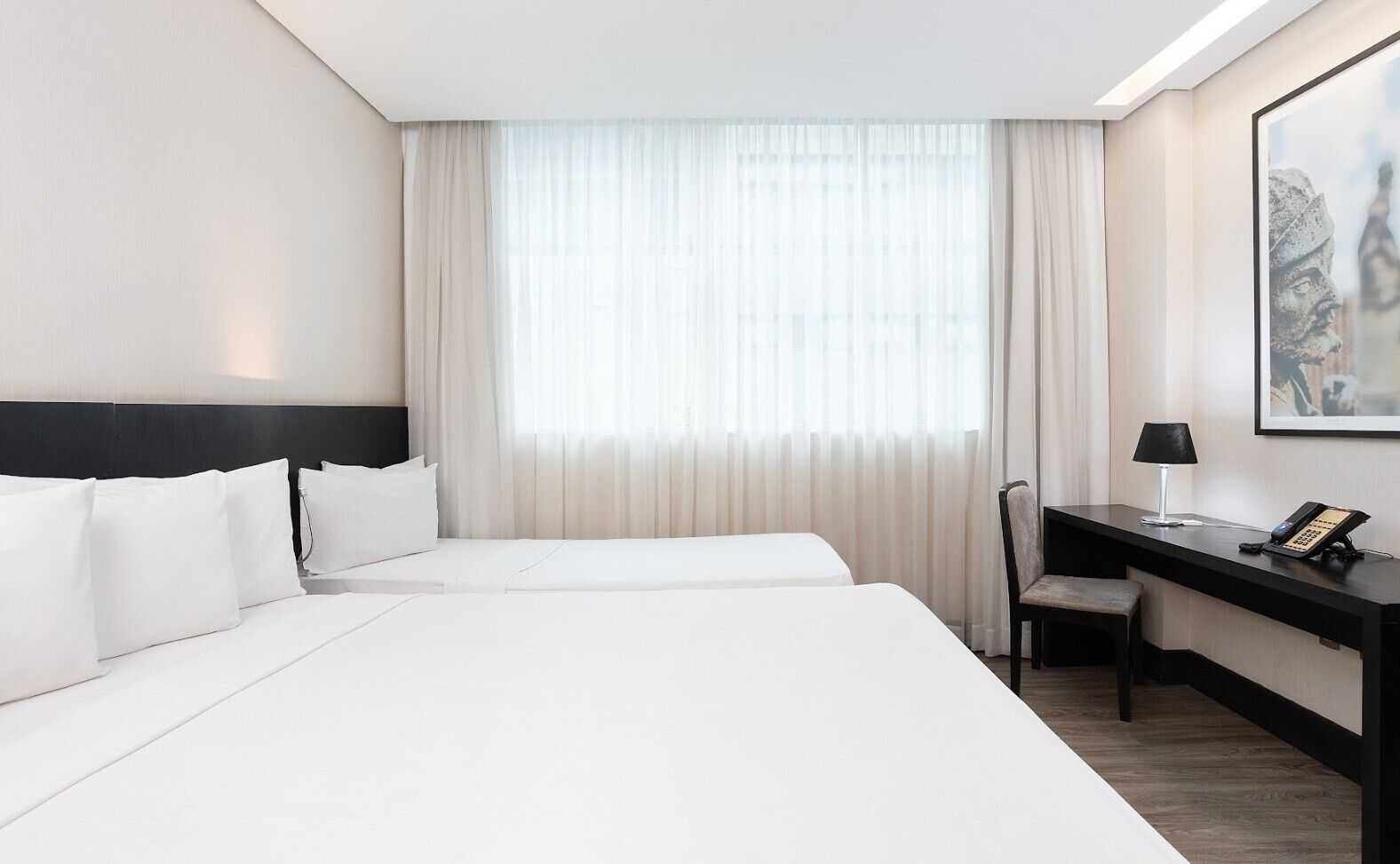 Habitación de hotel con dos camas, un escritorio con silla, un teléfono y una ventana con cortinas traslúcidas.