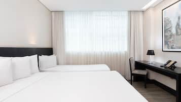 Habitación de hotel con dos camas, un escritorio con silla, un teléfono y una ventana con cortinas traslúcidas.