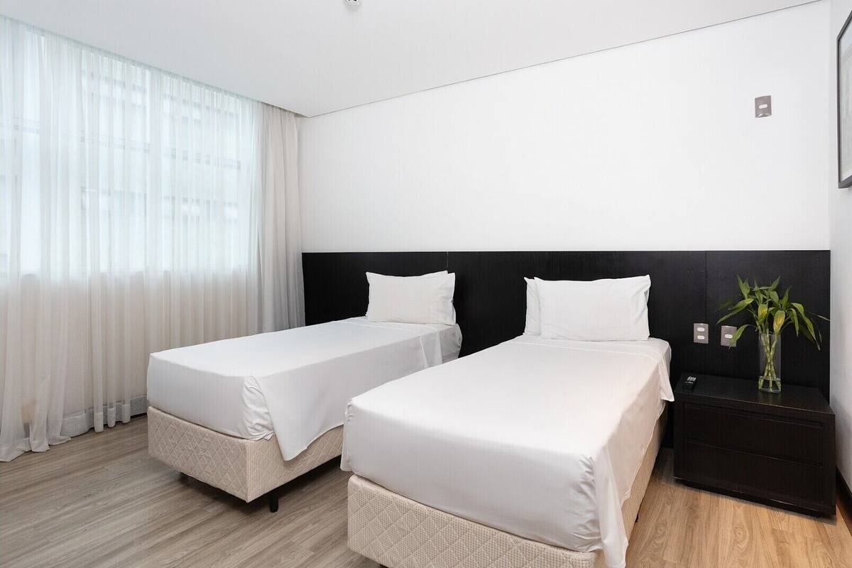 Habitación de hotel con dos camas individuales, cabecera oscura, una mesita de noche con un florero y cortinas de gasa.