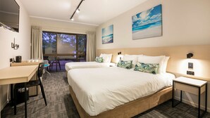 Ein Hotelzimmer mit zwei Betten, einem Schreibtisch und einem Sessel. Es gibt ein großes Fenster mit Vorhängen, einen an der Wand montierten Flachbildfernseher und Kunstwerke über den Betten.
