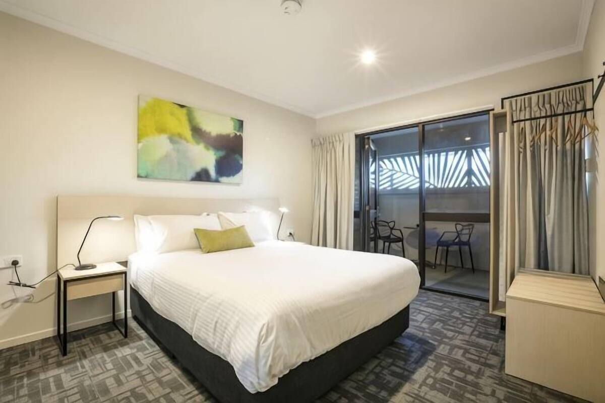 Ein Hotelzimmer mit einem großen Bett, einem Nachttisch mit Lampe, einem Sessel und Blick durch eine Glastür nach draußen.