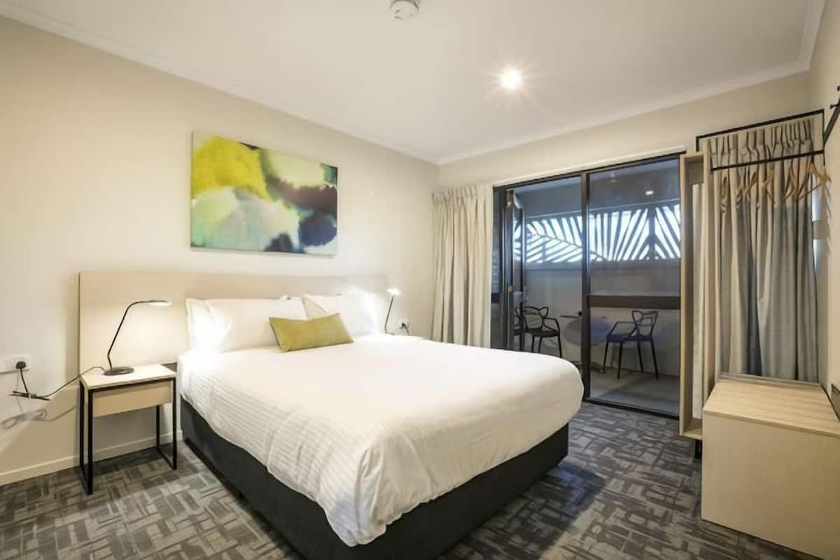 A hotel room with a large bed, a bedside table with a lamp, a chair, and a view of the outside through a glass door.