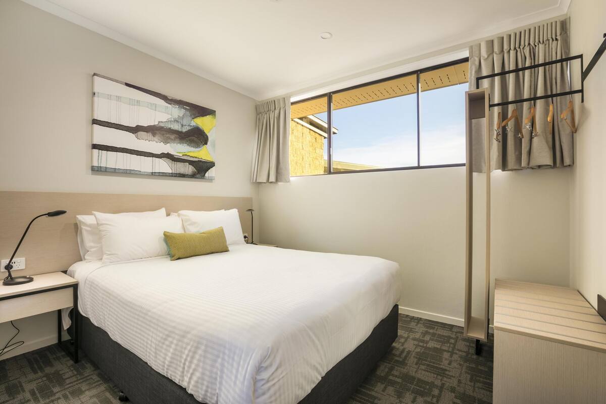 Ein Hotelzimmer mit einem großen Bett, einem Schreibtisch mit Lampe, einem Fenster mit Vorhängen und einem Wandhaken für Kleidung.