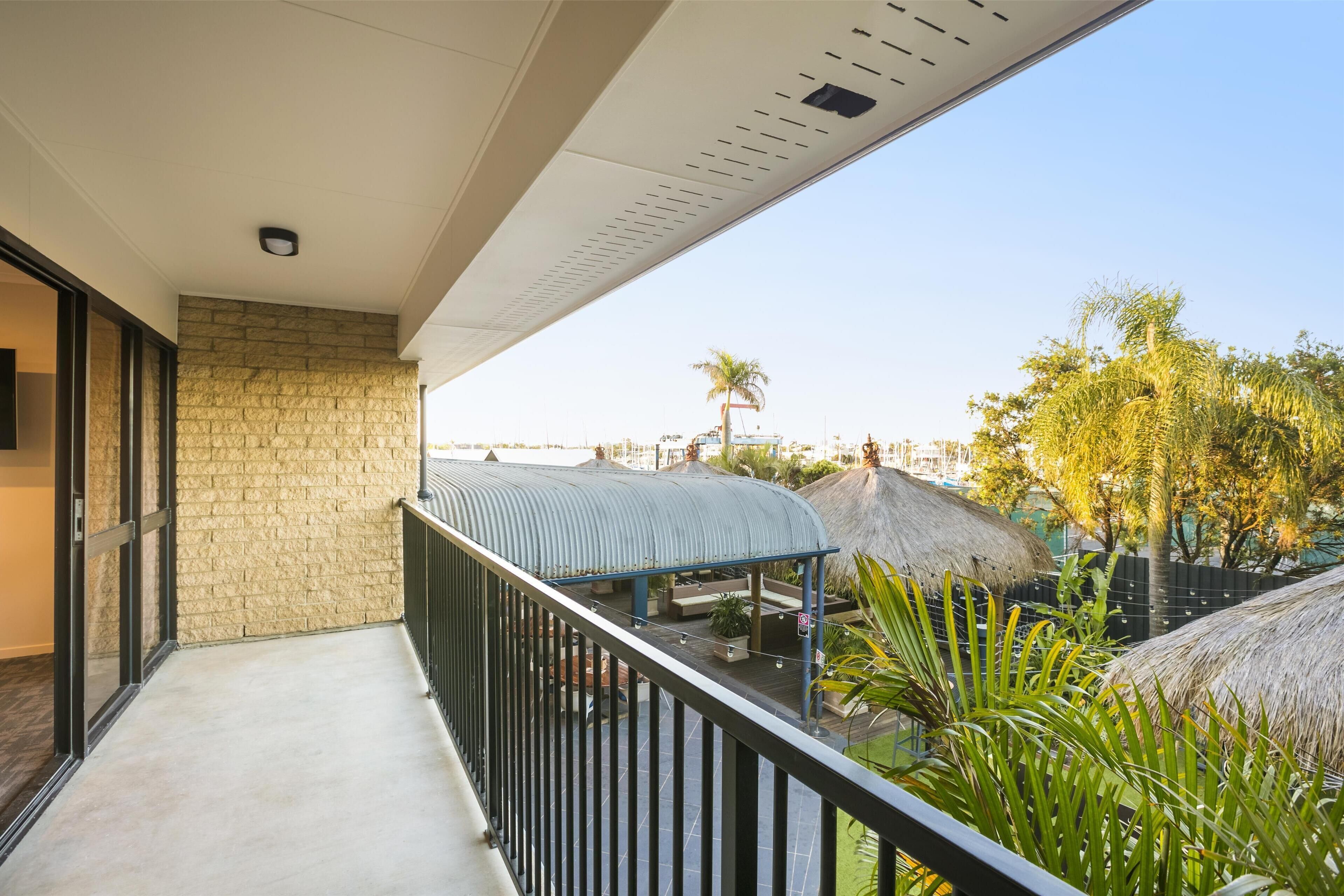 A balcony with a stone wall, a metal railing, and a view of a resort area with thatched umbrellas and palm trees.