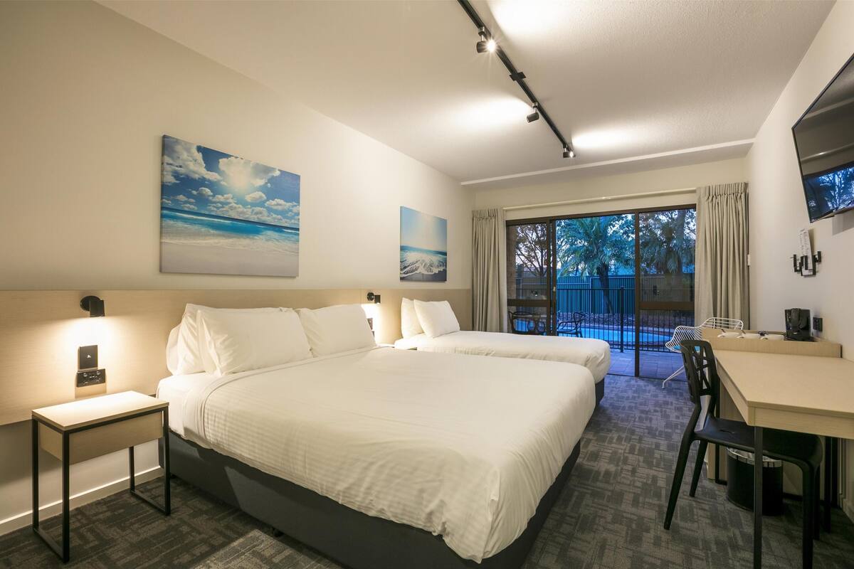 Ein Hotelzimmer mit einem großen Bett, einem Schreibtisch, einem Sessel und einem Fernseher. Es gibt ein Fenster mit Blick auf Palmen und einen Balkon.