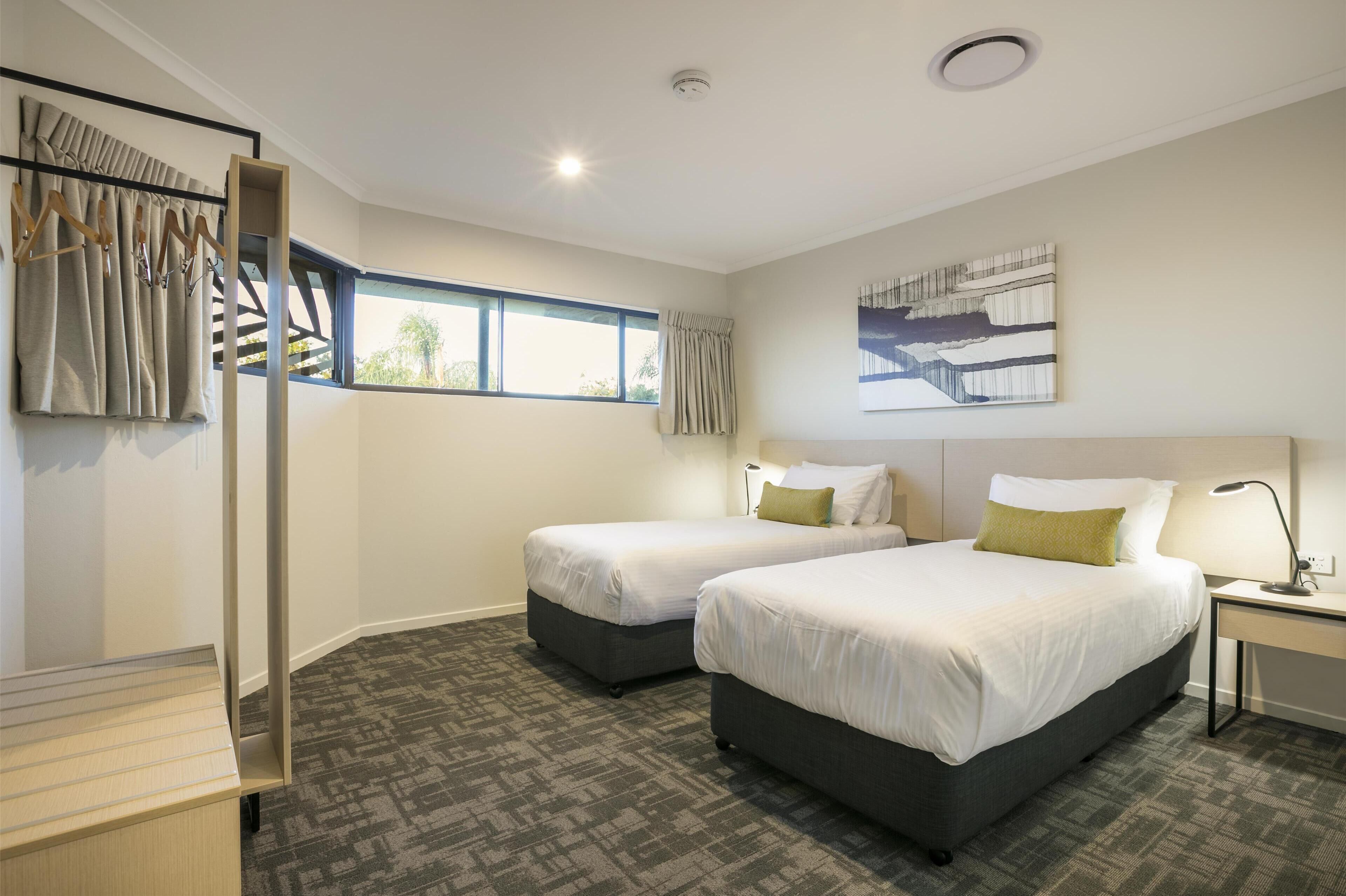 A hotel room with two beds, a desk, a chair, a window with curtains, a ceiling light, and a painting on the wall.
