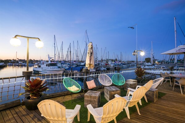 Un puerto deportivo con barcos amarrados, asientos exteriores con sillas y mesas, y luces colgantes.