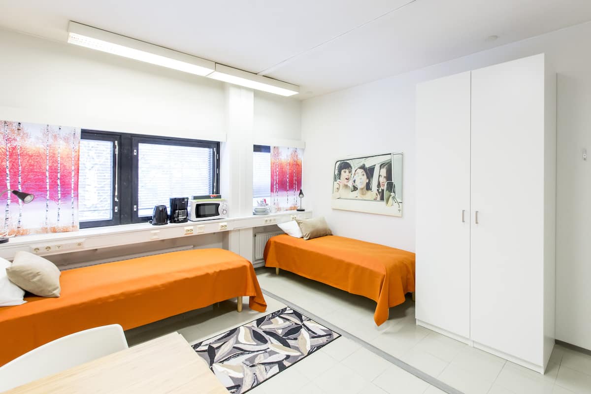 A room with two single beds, a desk with a computer, a microwave, and a coffee maker. There is a large wardrobe and a window with curtains.