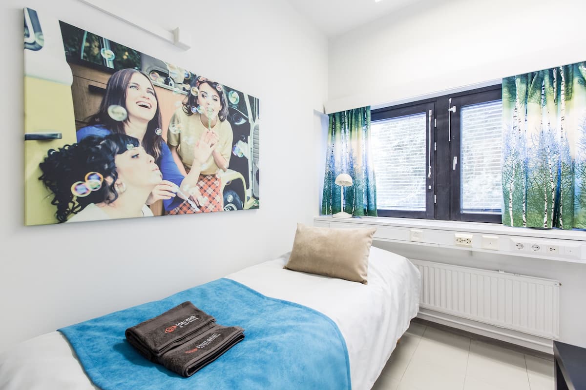 A bedroom with a bed, a blue blanket, a pillow, a window with curtains, and a painting on the wall.