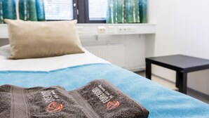 A bed with a blue blanket and two towels with a logo, a pillow, a window with curtains, and a small black table. - Forenom Hostel Vantaa Airport (Vantaa)