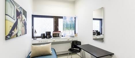 Una habitación pequeña y funcional con una cama individual, un escritorio, una silla, un cubo de basura negro y una ventana con persianas.