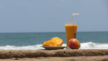 A glass of orange juice with a straw, a halved mango, and a whole mango on a plate, set against a beach backdrop with the ocean and a clear sky.