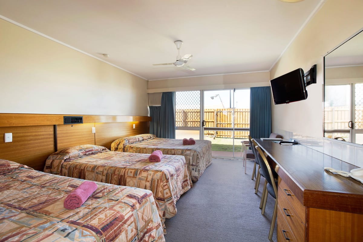 A hotel room with three beds, a wooden desk, a flat-screen TV, and a balcony with a view.
