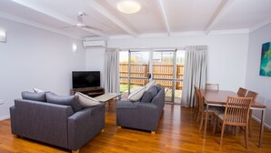 A modern living room with a flat-screen TV, grey sofas, wooden flooring, a dining table with chairs, and a large sliding glass door leading to an outdoor area. - Wilsonton Hotel (Wilsonton)