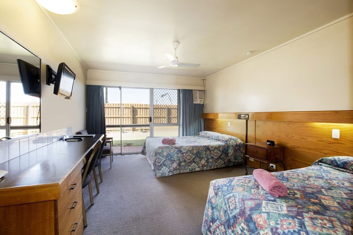 A room with a desk, two beds, a ceiling fan, and a view of the outside.