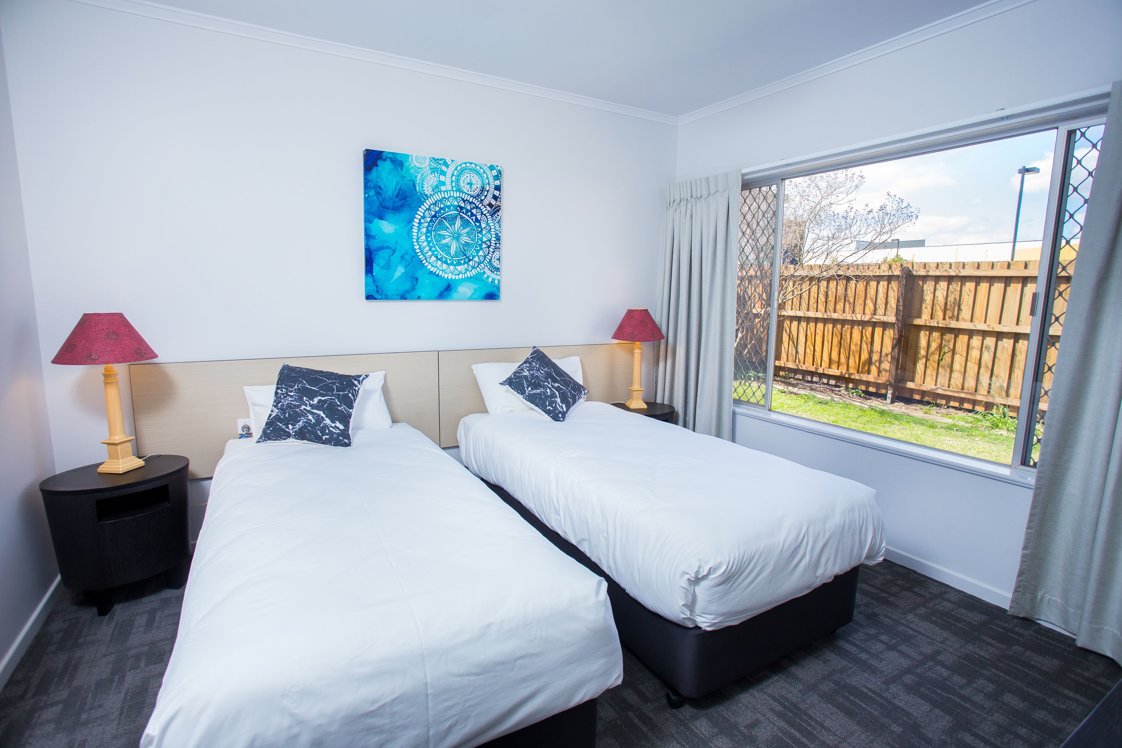 Habitación de hotel con dos camas, un ventanal con vista a una cerca de madera y un cuadro abstracto azul en la pared.