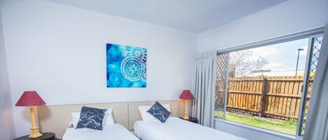 Habitación de hotel con dos camas, un ventanal con vista a una cerca de madera y un cuadro abstracto azul en la pared.