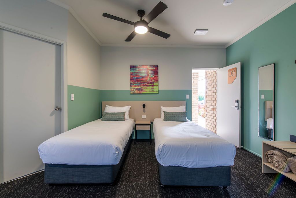 Habitación de hotel con dos camas individuales, ventilador de techo, un cuadro, pared de ladrillo y una puerta.