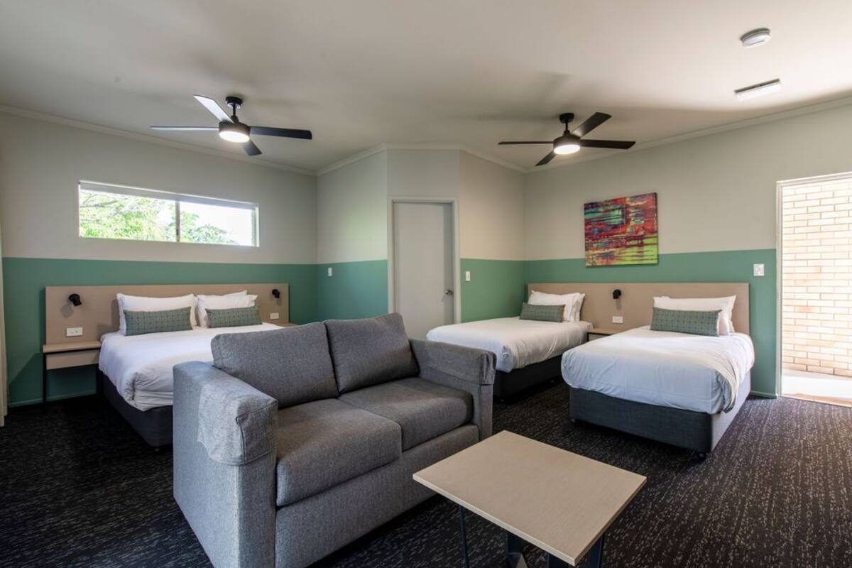 Ein Hotelzimmer mit zwei Einzelbetten, einem Schlafsofa, einem kleinen Tisch und zwei Deckenventilatoren.