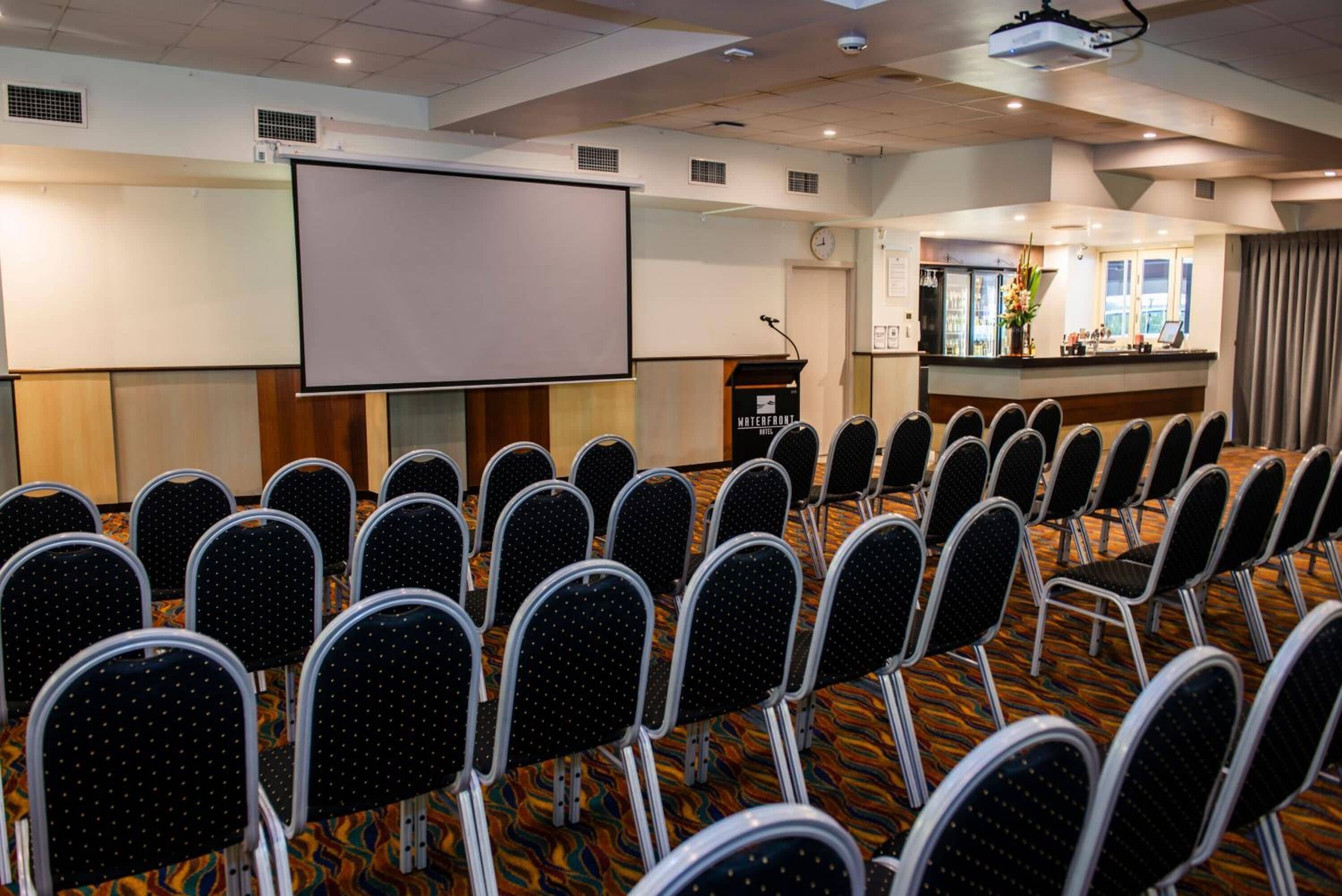 Een vergaderzaal met rijen stoelen, een groot scherm en een spreekgestoelte.