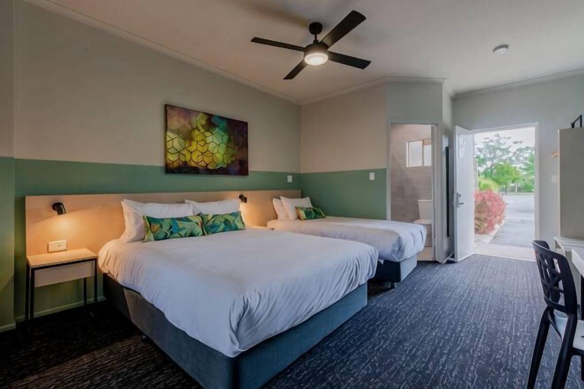 Ein Hotelzimmer mit zwei Betten, einem Deckenventilator, einem Gemälde und Blick ins Freie.