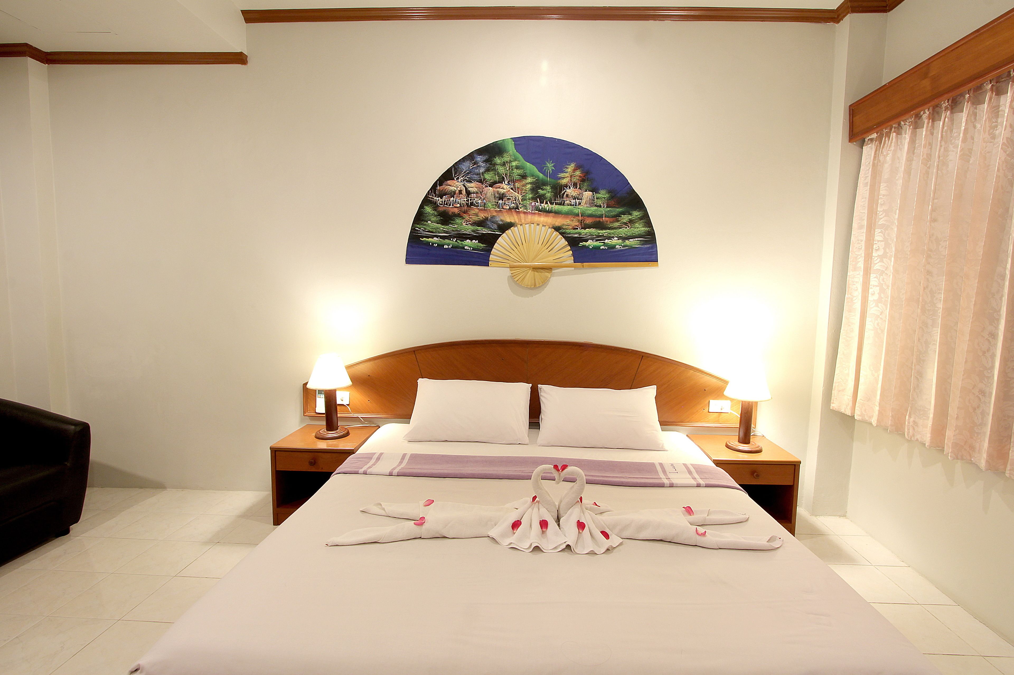 A hotel room with a bed, two bedside lamps, a headboard, a wall-mounted painting, and a window with curtains.