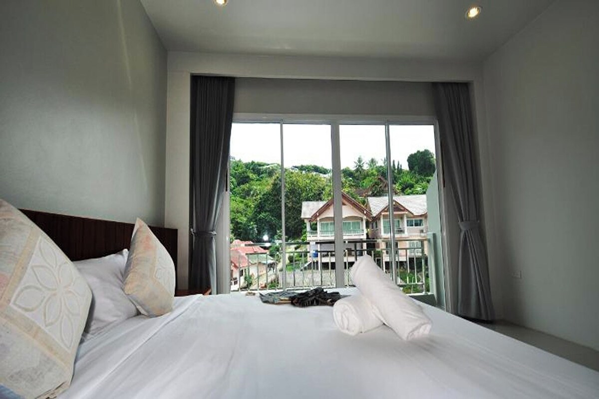A hotel room with a large bed, a view of a building through a window, and a balcony.