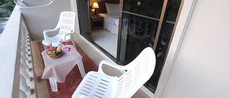 A balcony with two white plastic chairs, a small table with a basket of fruit and a glass of wine, and a nightstand with a lamp.