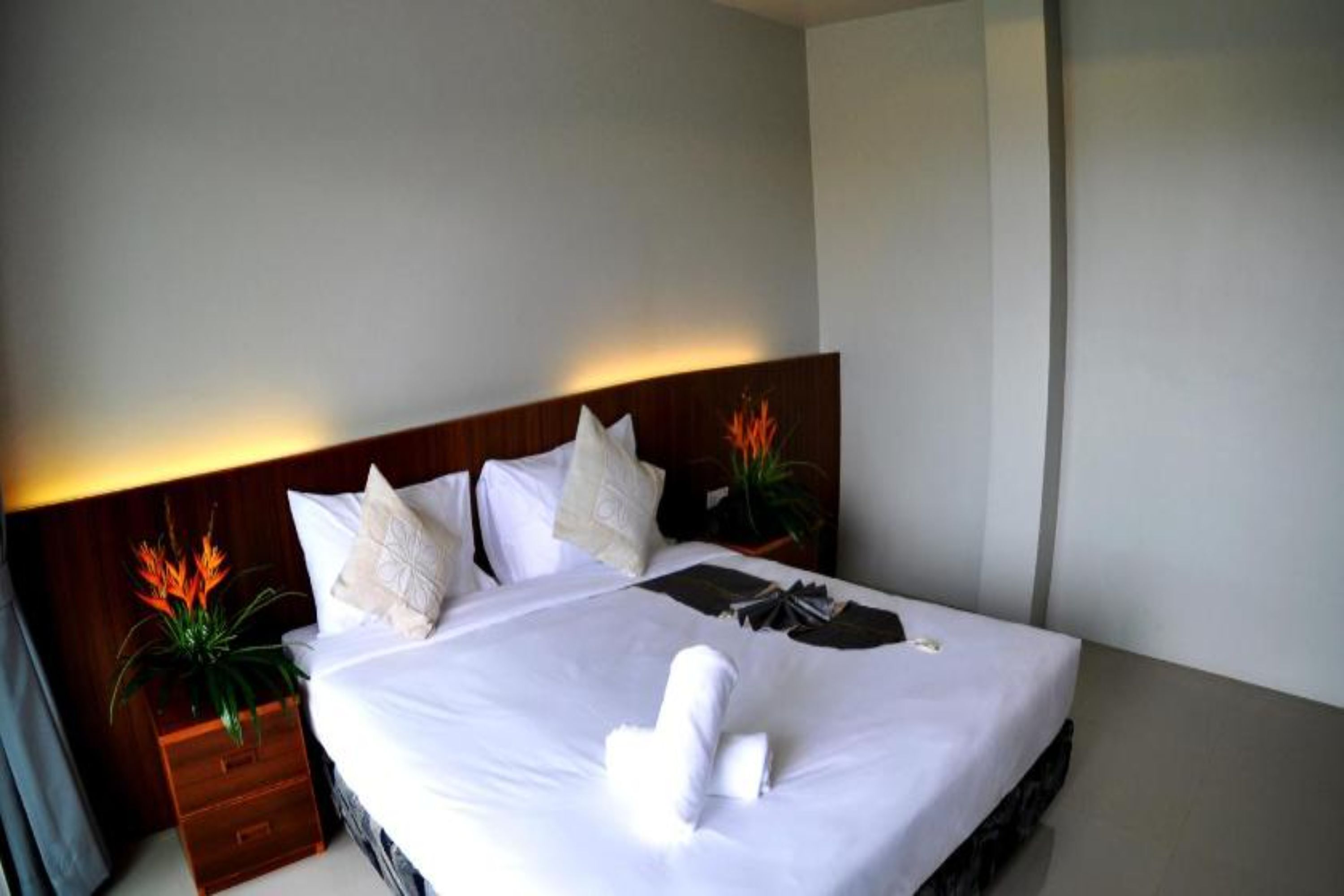 Una cama bien hecha con sábanas blancas, una toalla doblada y una almohada decorativa. Hay una mesita de noche de madera con una planta al lado de la cama.