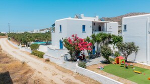 A coastal building with a white exterior, a small garden with pink flowers, and a play area with a red-roofed structure. - Diamantis Studios (Naxos)