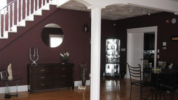 A spacious living room with a wooden floor, a staircase with white railings, a dark-colored sofa, and a dining area with a table and chairs. - Driftwood Heights Bed & Breakfast (Summerside)