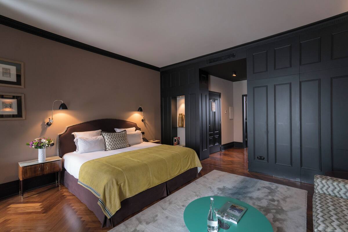 Una habitación de hotel con una cama grande, lámparas de noche, una mesita de noche, una manta verde, una mesa de centro redonda y una alfombra estampada.