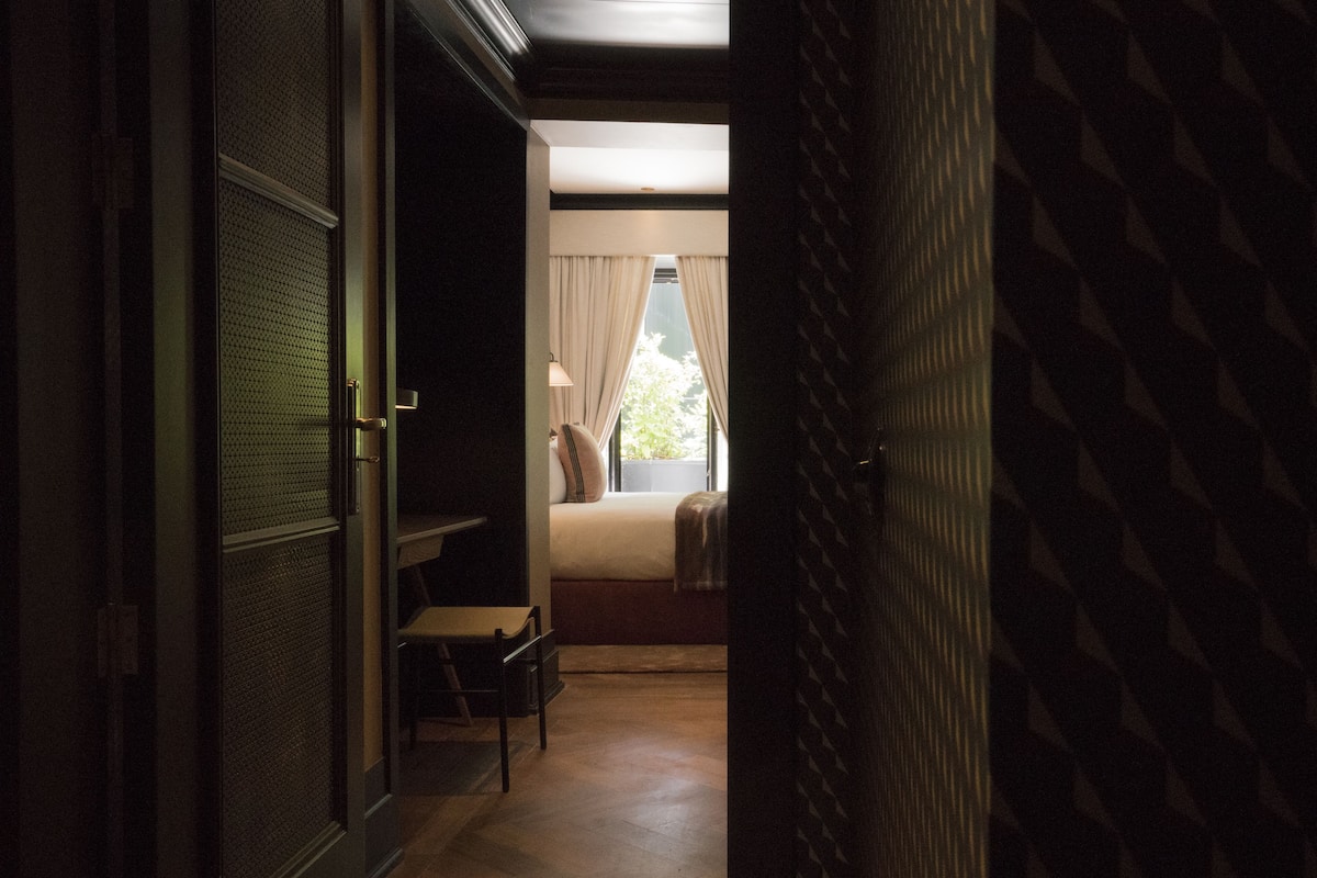 Un pasillo que conduce a un dormitorio con una cama, una silla y una ventana con cortinas.