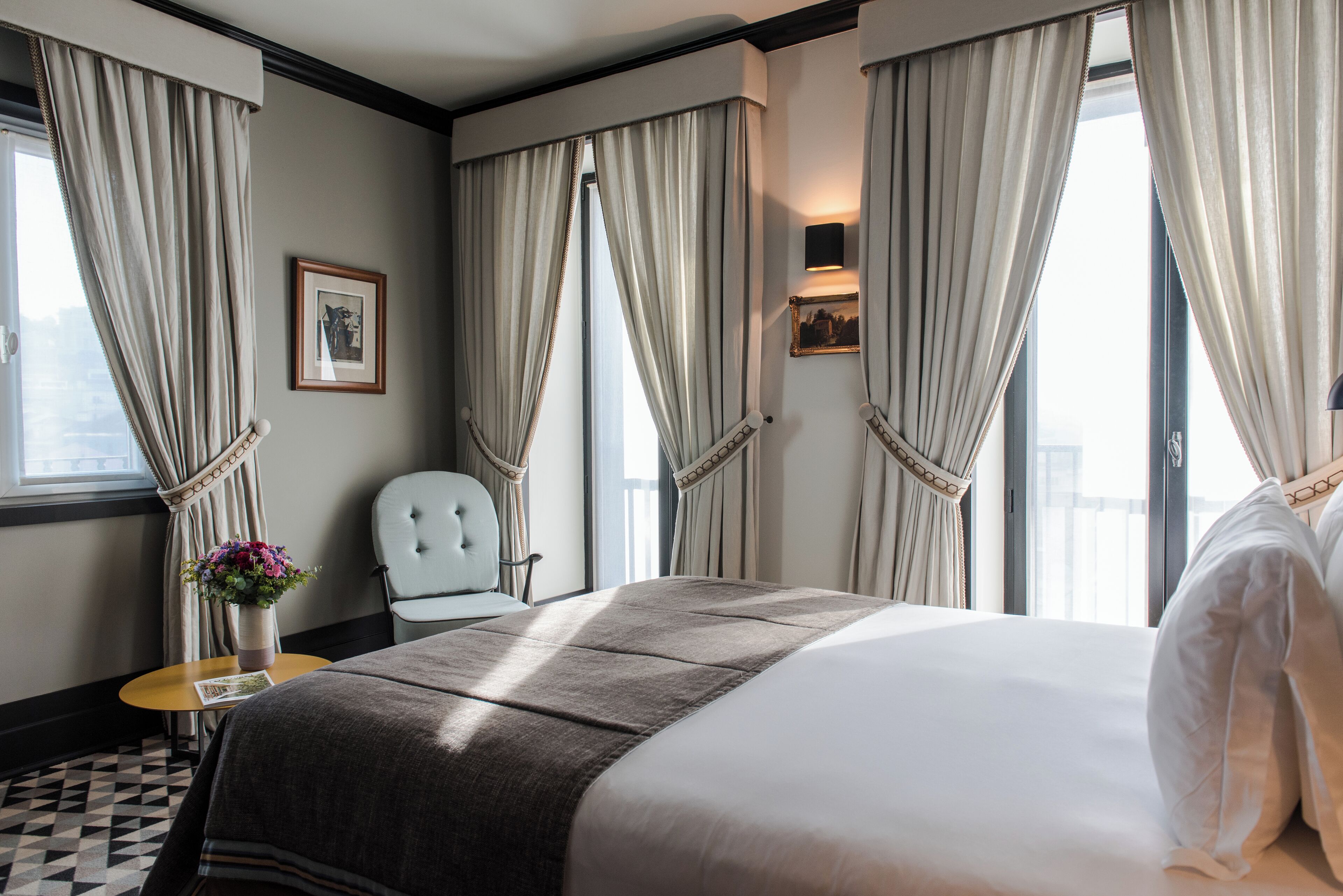 A hotel room with a large bed, a chair, a small table with a vase of flowers, and curtains on the windows.
