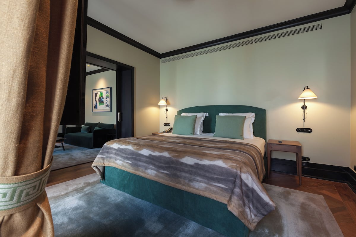 Habitación de hotel con cama, dos lámparas de noche, una mesita de noche, una pequeña mesa, un sofá y vistas al pasillo.