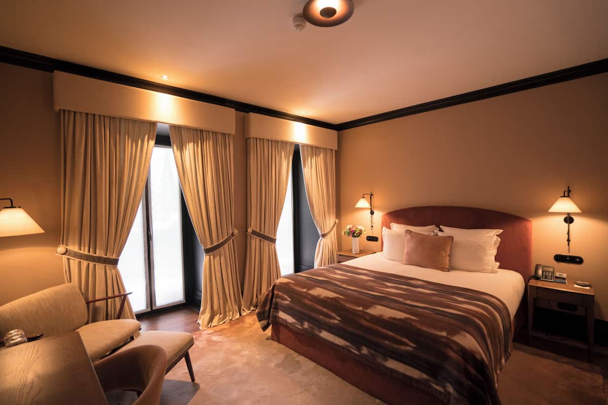 Una habitación de hotel con una cama grande, dos lámparas de noche, una silla y cortinas.