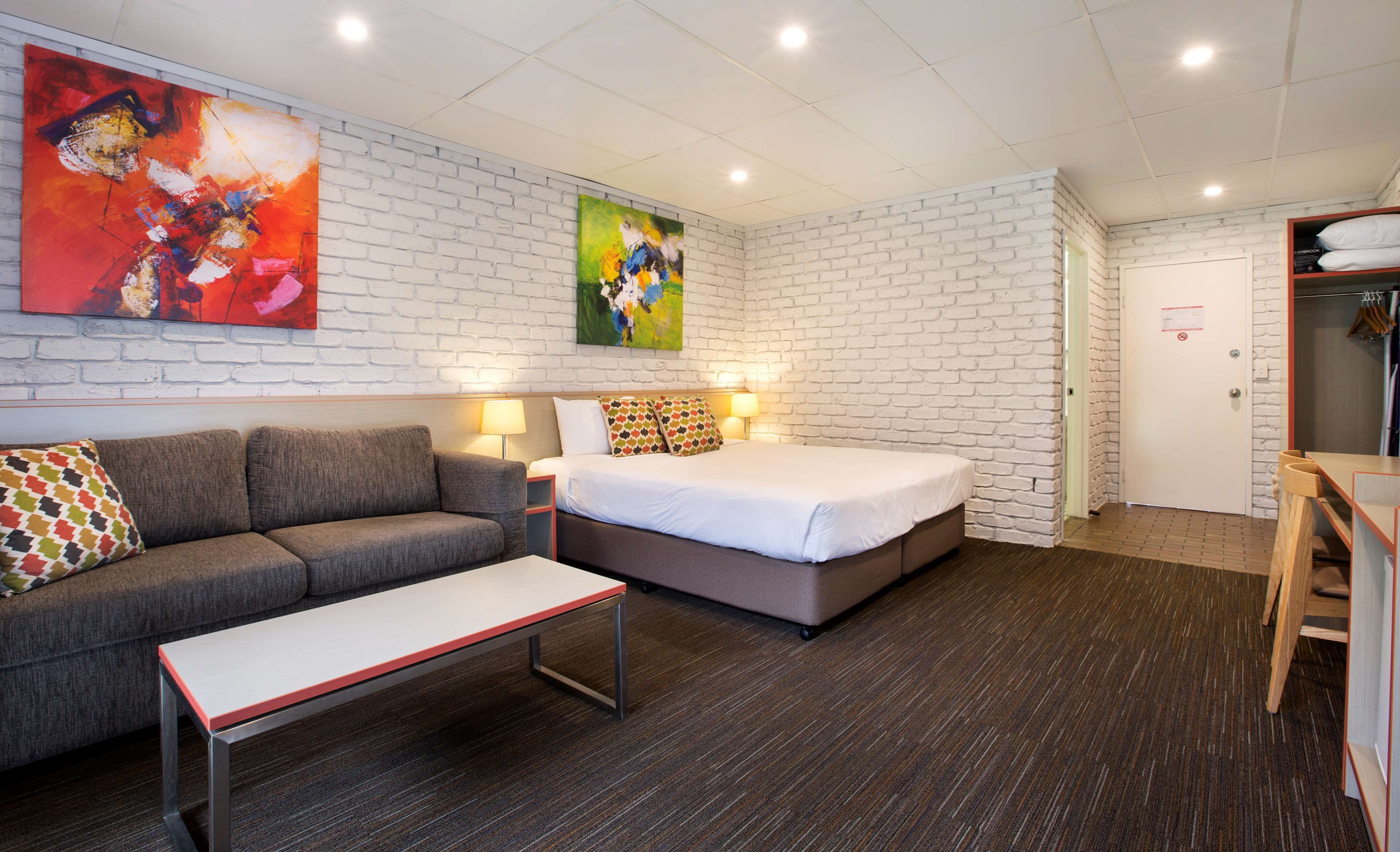 A hotel room with a white brick wall, a bed with colorful pillows, a gray sofa, a small table, and two bedside lamps.
