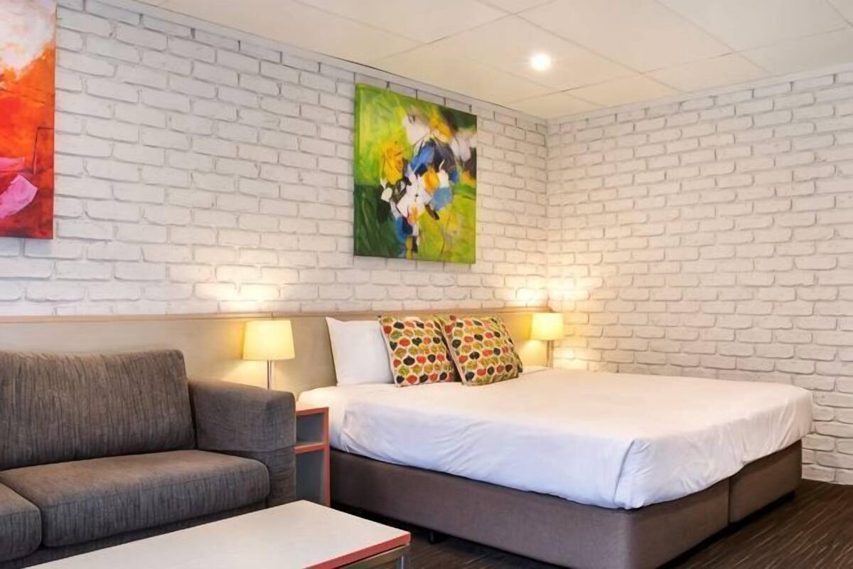 Een hotelkamer met een witte bakstenen muur, een bed met wit beddengoed, een grijze bank en twee wandlampen.