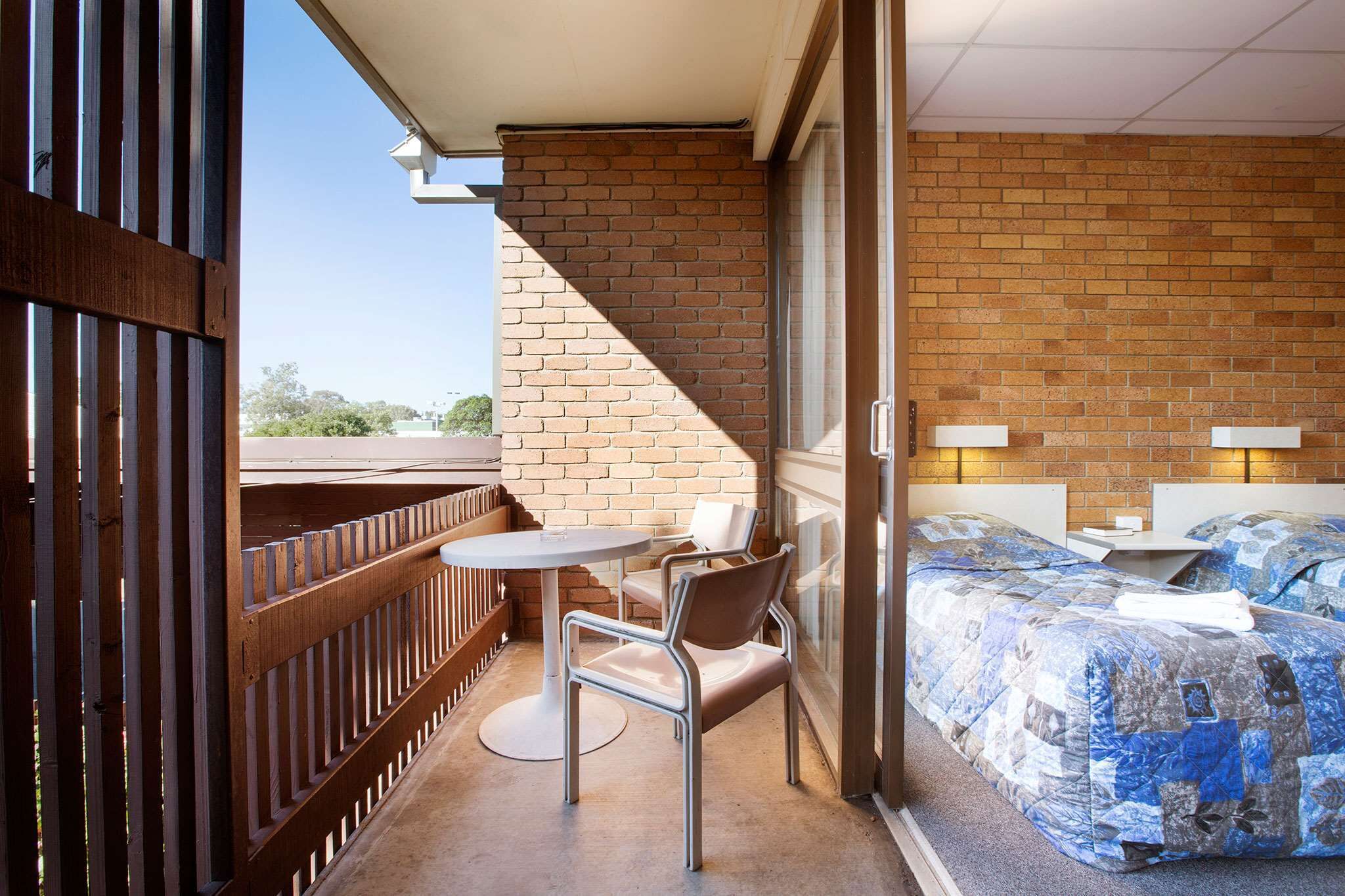 A balcony with a table and chairs, a bed with a blue quilt, and a brick wall.