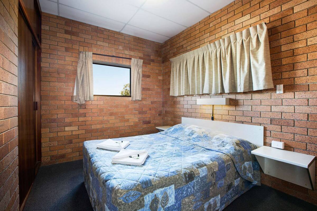 Een kleine eenpersoonskamer met een bakstenen muur, een raam met gordijnen, een nachtkastje en een wandplank.