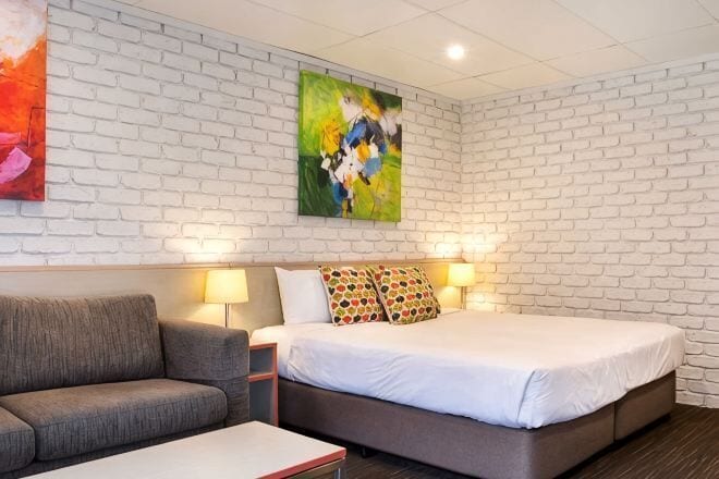 Hotellihuone, jossa on valkoisesta tiilestä tehty seinä, valkoisilla lakanoiden peitetty sänky, harmaa sohva ja kaksi seinälle asennettua lamppua.
