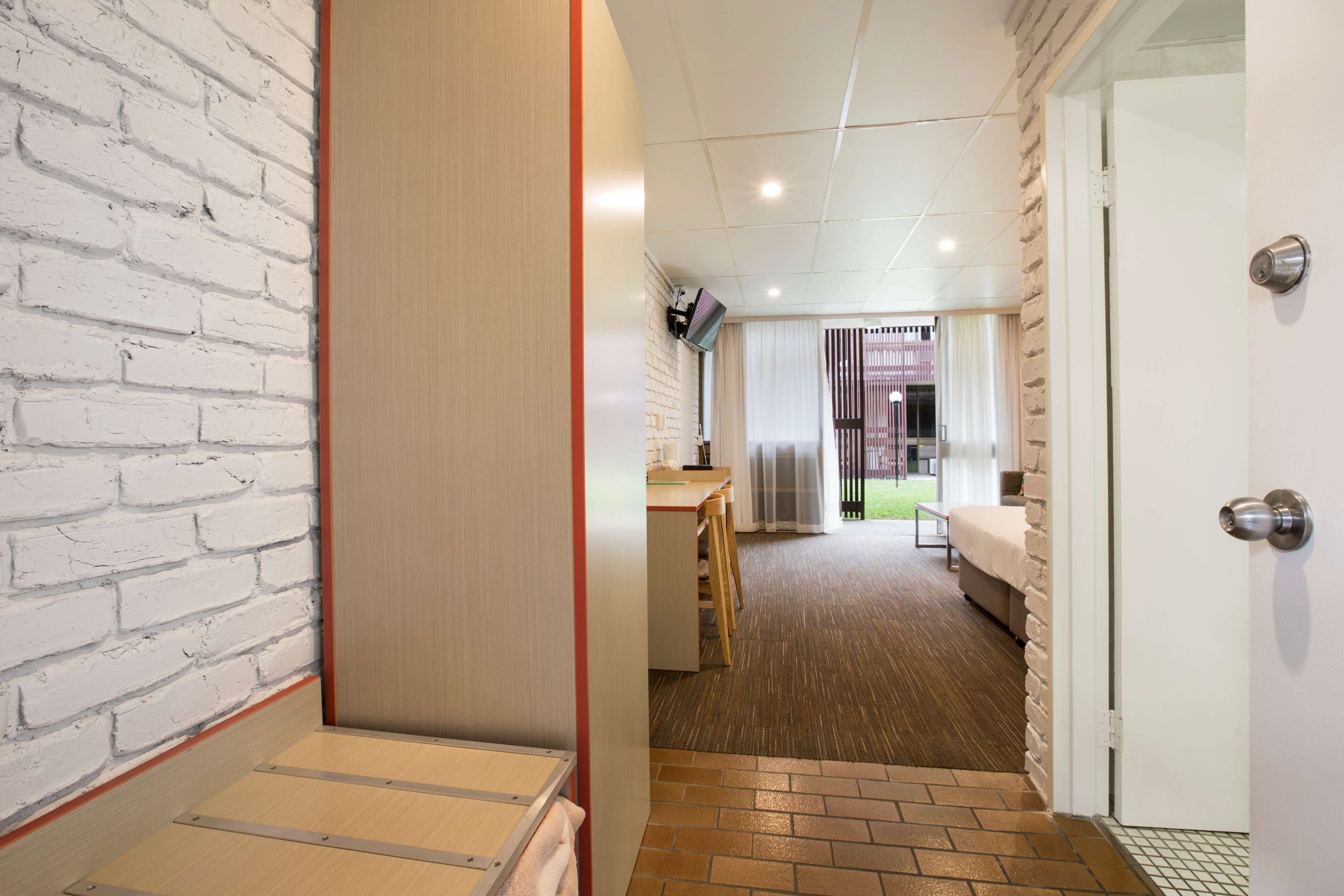 Un couloir d’hôtel avec un mur en briques blanches, une porte en bois et un sol carrelé, menant à une chambre dotée d’un lit et d’une fenêtre.