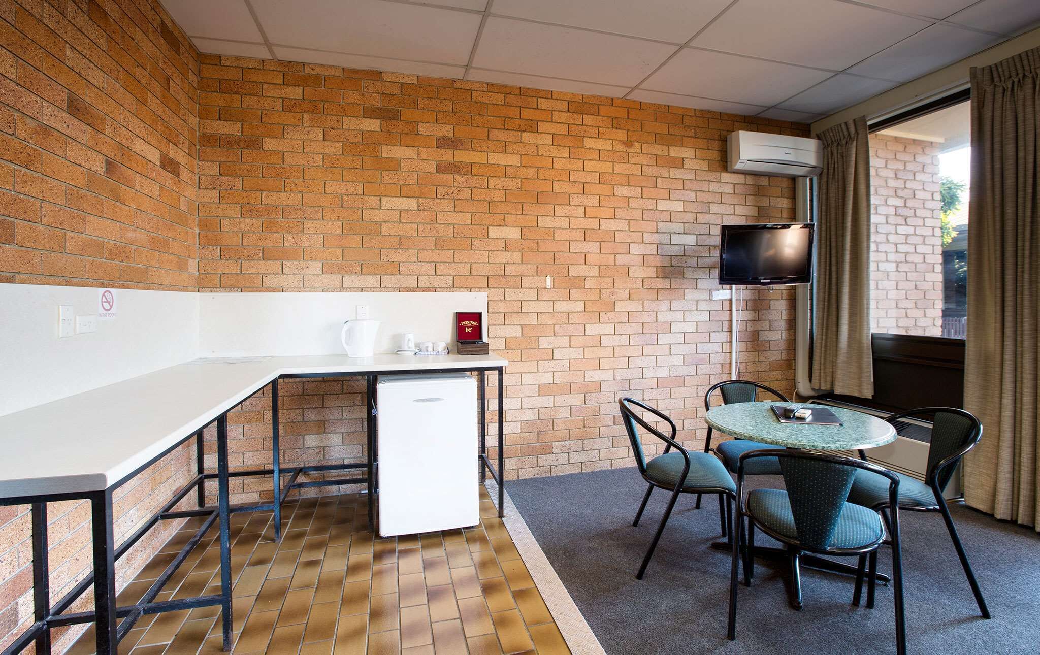 Une petite pièce moderne avec un mur en briques, un bureau blanc, un petit réfrigérateur, une télévision et une table ronde avec des chaises.