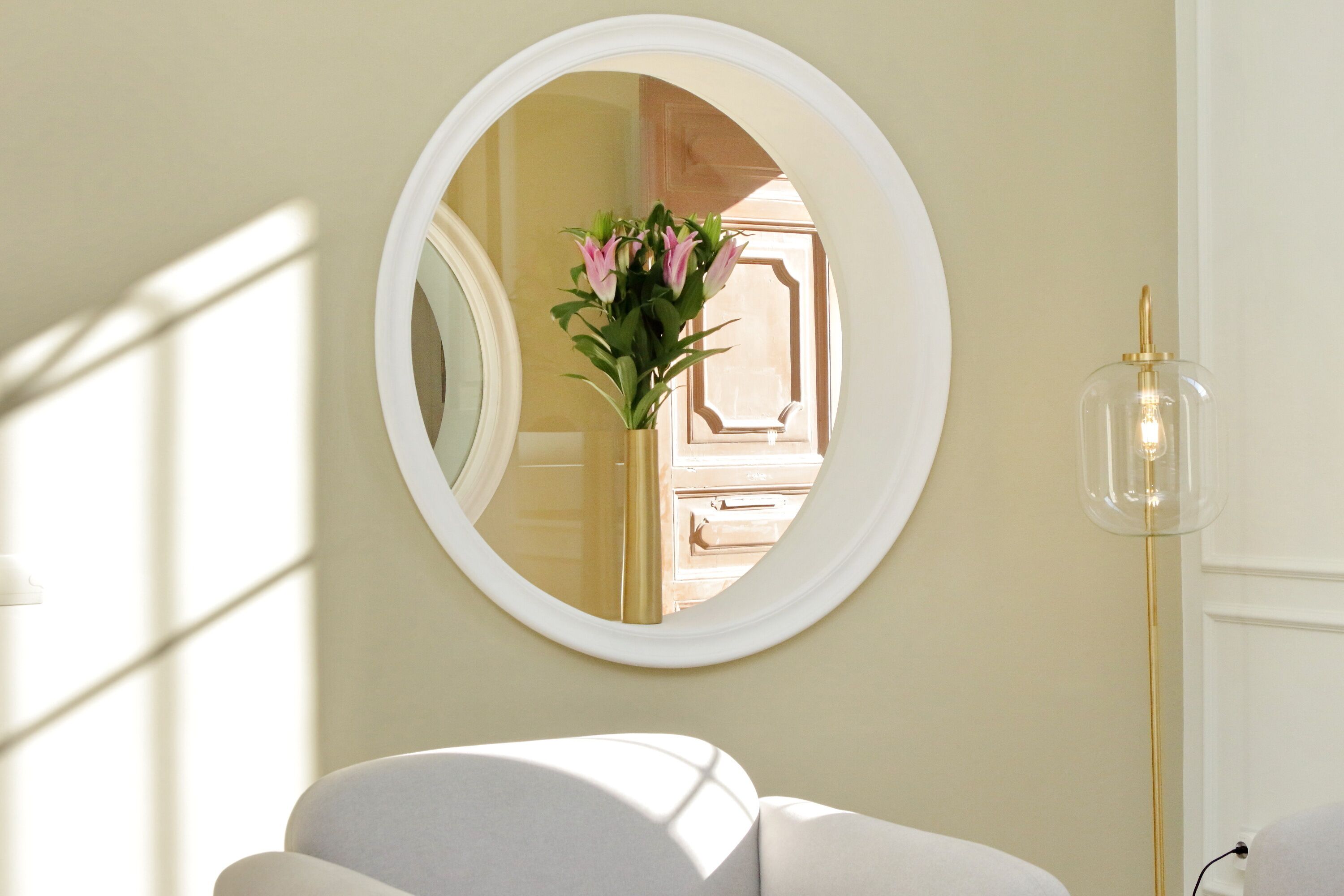 A round mirror on a wall, a white chair, and a tall floor lamp.