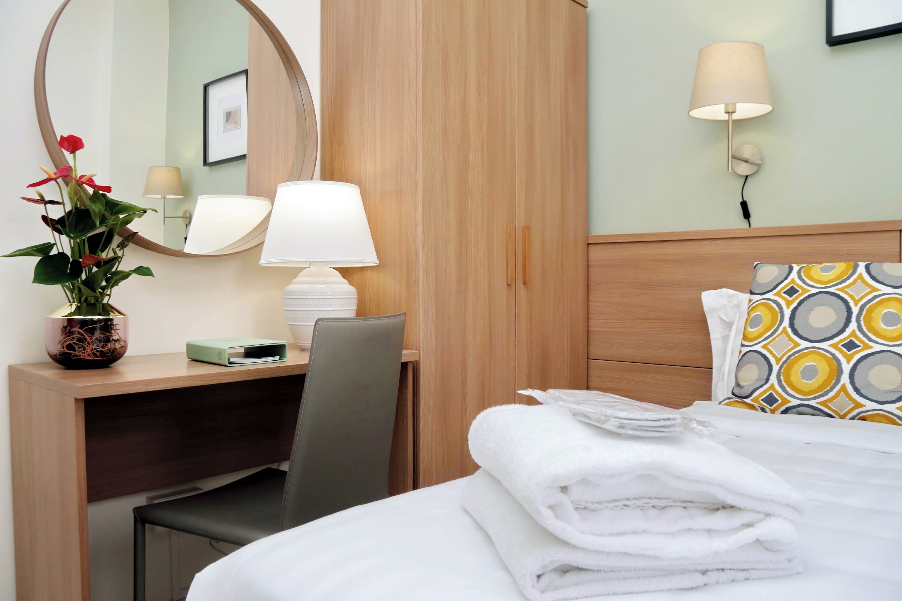 Une chambre d’hôtel équipée d’un lit, d’un bureau, d’une chaise, d’une lampe, d’un miroir et d’une plante en pot.